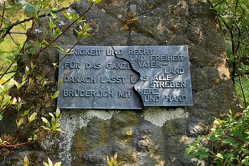 Farbfoto: In der Mitte gebrochene Steintafel mit der Aufschrift: "Einigkeit und Recht und Freiheit für das ganze Vaterland, danach lasst uns alle streben, brüderlich mit Herz und Hand."